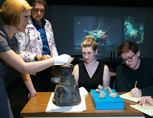 Professor and students studying Pre-Columbian terracotta whistles and a 19th century wooden helmet mask from West Africa.